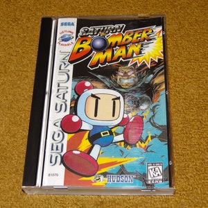 May include: A Sega Saturn video game case with a blue and red cover featuring the title "Saturn Bomber Man". The cover art shows a cartoon character with a white head and red gloves, kicking a yellow bomb. The game is rated "KA" for Kids to Adults.