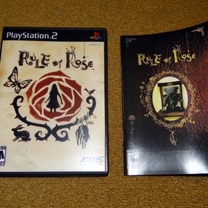 May include: A PlayStation 2 video game case and instruction manual for the game "Rule of Rose". The game case features a black and red rose with a silhouette of a girl in the center. The instruction manual has a black background with a gold-colored rose and a teddy bear in a cage.