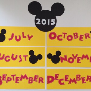 May include: A set of six yellow cards with red Mickey Mouse-shaped letters spelling out the months of July, August, September, October, November, and December. Each card has a black Mickey Mouse head on the side. The top card has a black Mickey Mouse head with the year 2015 in white text.