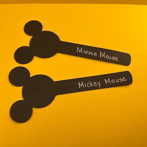 May include: Two black paper cutouts shaped like Mickey Mouse heads with extended rectangular handles. The top cutout has the white text Minnie Mouse written on the handle, and the bottom cutout has the white text Mickey Mouse written on the handle.