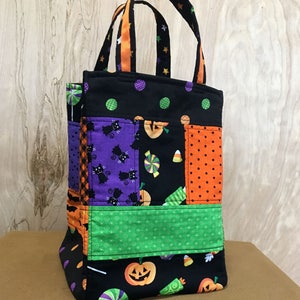 May include: A black Halloween trick-or-treat bag with a patchwork design featuring black cats, pumpkins, candy, and polka dots. The bag has a green fabric pocket and orange ribbon handles.