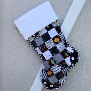 May include: A handmade Christmas stocking with a patchwork design. The stocking features a white cuff and is adorned with gold bells. The body is composed of black, white, and gray squares with festive patterns, and a black and white polka dot and striped border.