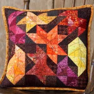 Puede incluir: Una almohada decorativa con un diseño de patchwork que presenta hojas de otoño en tonos de naranja, amarillo, rojo y marrón. La almohada tiene un borde de terciopelo marrón.