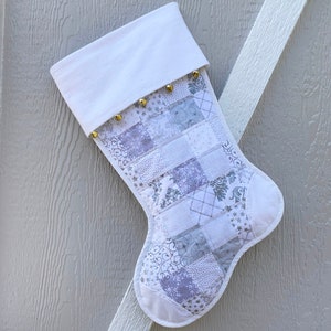 Puede incluir: Calcetín de Navidad blanco con un diseño de patchwork de tela blanca y plateada. El calcetín tiene un puño blanco y cascabeles dorados.