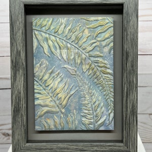 May include: A framed artwork featuring a textured, embossed design of fern leaves in shades of gold and green on a blue background.