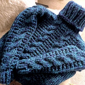 Könnte beinhalten: Ein grob gestrickter Pullover in Türkis- und Blautönen. Der Pullover hat einen gerippten Rollkragen und ein strukturiertes Zopfmuster. Das dicke Garn erzeugt ein gemütliches und warmes Aussehen. Der gefaltete Pullover wird auf einer neutralen Oberfläche präsentiert.
