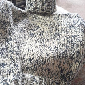 Könnte beinhalten: Ein grob gestrickter Pullover in Creme und Marineblau. Er hat einen hohen, gerippten Rollkragen und ein dickes, strukturiertes Strickmuster. Das Design ist gemütlich und warm, ideal für kühlere Temperaturen. Ein klassisches, bequemes Kleidungsstück.