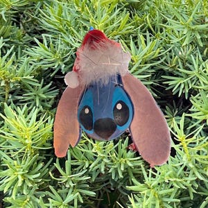 May include: A blue and black character ornament with large pink ears and a Santa hat. The ornament is set against a backdrop of green foliage. The character has large black eyes and a black nose.