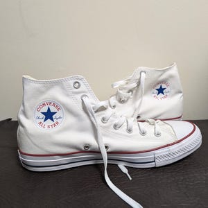 Può includere: Un paio di scarpe alte in tela bianca con lacci bianchi, occhielli in metallo argentato e punta in gomma. Il lato presenta una toppa circolare Converse Chuck Taylor All Star con una stella blu e una suola con strisce rosse e blu.