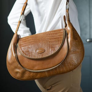May include: A brown leather shoulder bag with a crocodile-embossed design. The bag has a flap closure and a zipped pocket on the front. The bag is being worn by a person with a white shirt.