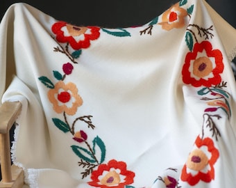 May include: An off-white throw blanket featuring a vibrant floral embroidery pattern in shades of red, orange, and dark green. The blanket has a textured fabric surface and frayed edges along the sides, creating a soft and casual appearance.