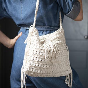 Op de afbeelding: Crèmekleurige macramé schoudertas met franjedetails. De tas heeft een geweven ontwerp en een gevlochten band. De tas wordt over de schouder gedragen. De tas is gemaakt van een zacht, geweven materiaal.