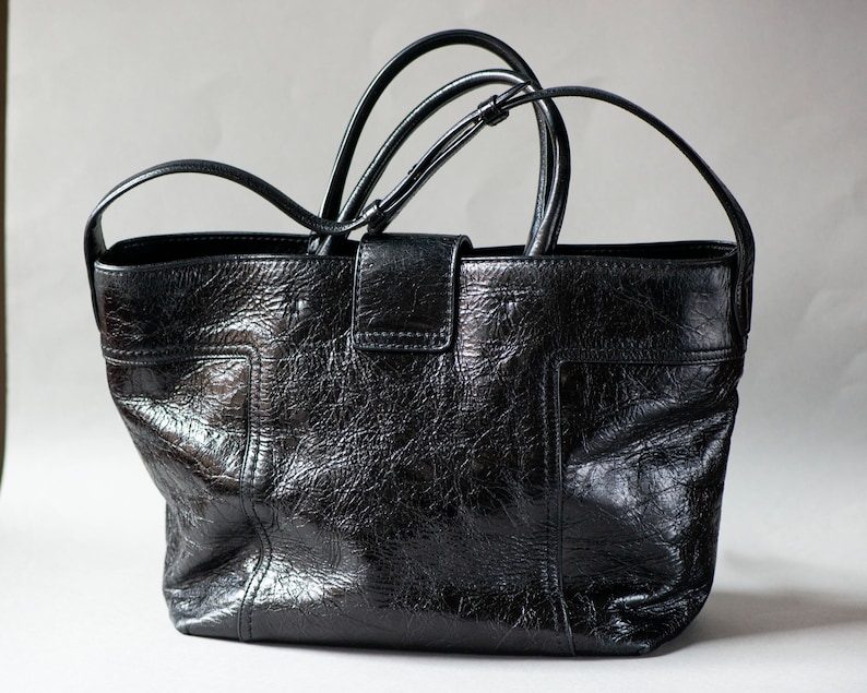 Un sac cabas Tod's en cuir noir avec fermeture à boucle argentée. Ce sac en cuir texturé est doté d'une bandoulière longue. Il présente un fermoir à boucle orné du logo Double T ton sur ton sur la languette avant. Une bandoulière plate et longue en cuir est également fournie pour un porté épaule polyvalent.