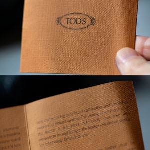 Op de afbeelding: Een close-up van een bruin lederen verzorgingsetiket met het TOD'S-logo en instructies voor het verzorgen van het leer. De tekst luidt: "Artikel vervaardigd van hoogwaardig geselecteerd kalfsleer en gelooid om de natuurlijke eigenschappen te behouden. De nerven, die typerend zijn voor dit leer, worden met opzet intact gelaten, met de tijd kan het leer door blootstelling aan lucht en zonlicht licht donkerder worden. Krassen ontstaan gemakkelijk. Fijne leer. In geval van contact met water moet het leer worden beschermd en onmiddellijk met een zachte doek worden gedroogd om licht opzwellen of watervlekken te voorkomen."