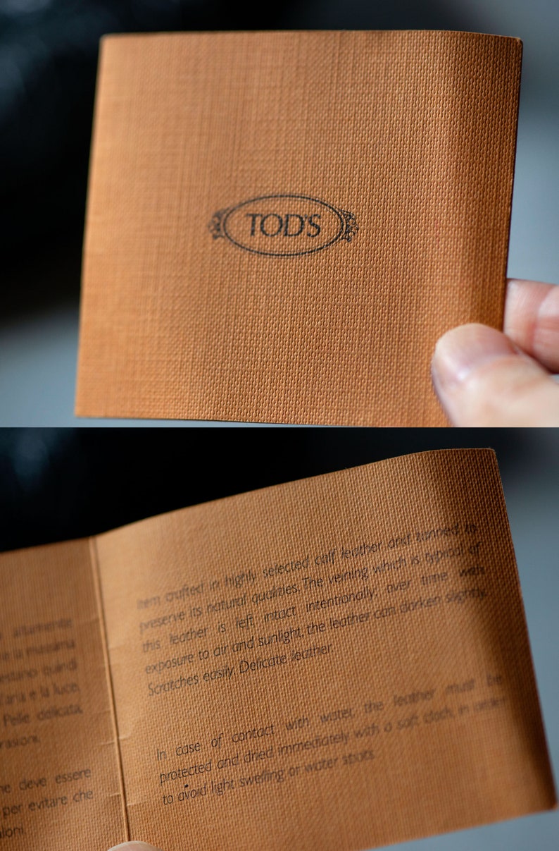 Op de afbeelding: Een close-up van een bruin lederen verzorgingsetiket met het TOD'S-logo en instructies voor het verzorgen van het leer. De tekst luidt: "Artikel vervaardigd van hoogwaardig geselecteerd kalfsleer en gelooid om de natuurlijke eigenschappen te behouden. De nerven, die typerend zijn voor dit leer, worden met opzet intact gelaten, met de tijd kan het leer door blootstelling aan lucht en zonlicht licht donkerder worden. Krassen ontstaan gemakkelijk. Fijne leer. In geval van contact met water moet het leer worden beschermd en onmiddellijk met een zachte doek worden gedroogd om licht opzwellen of watervlekken te voorkomen."