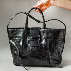 A black leather Tods tote bag with a silver T-shaped buckle closure. The bag has a structured shape and a top handle.
