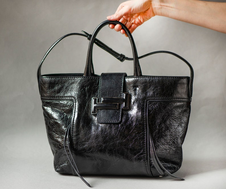 A black leather Tods tote bag with a silver T-shaped buckle closure. The bag has a structured shape and a top handle.