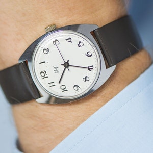 May include: A silver wristwatch with a white face and black leather band. The watch face has black Arabic numerals and a small second hand. The watch face also has the word "ОЛьфа" printed on it.