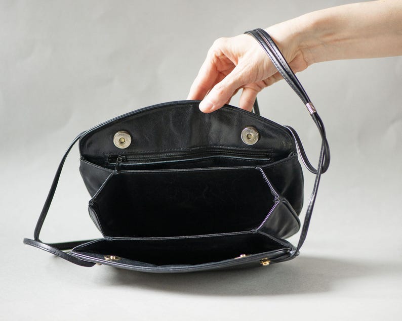 Puede incluir: Bolso de hombro de cuero negro con cierre de solapa y m&uacute;ltiples compartimentos. El bolso tiene una correa larga y delgada y est&aacute; abierto, revelando el interior. El bolso es sostenido por una mano.