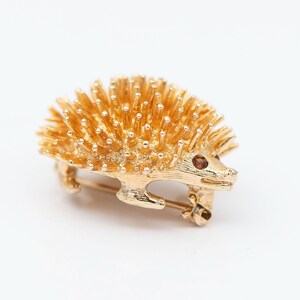 May include: A gold-toned brooch in the shape of a hedgehog. The hedgehog has a brown eye and is covered in small, gold-toned spikes.