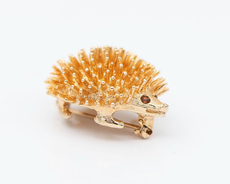 May include: A gold-toned brooch in the shape of a hedgehog. The hedgehog has a brown eye and is covered in small, gold-toned spikes.