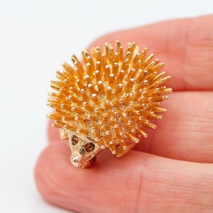 May include: A gold-toned brooch in the shape of a hedgehog. The hedgehog has a spiky back and two small brown eyes.