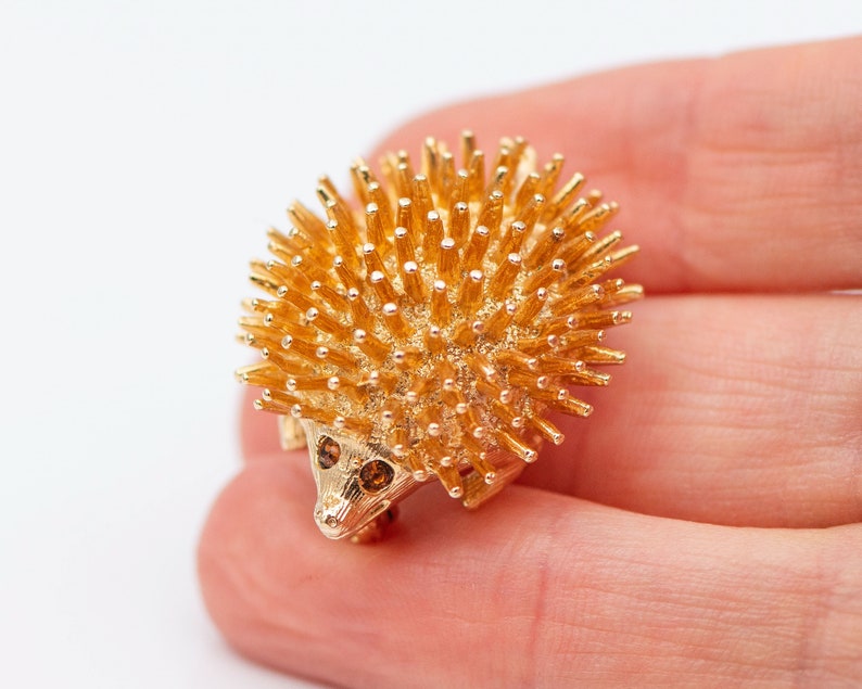 May include: A gold-toned brooch in the shape of a hedgehog. The hedgehog has a spiky back and two small brown eyes.