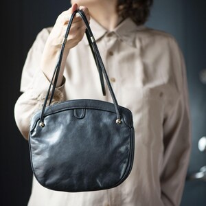 Puede incluir: Bolso de mano de cuero negro con forma redondeada y finas correas para el hombro. El bolso tiene detalles dorados en los laterales y un peque&ntilde;o cierre de solapa. La persona sostiene el bolso frente a un fondo neutro.