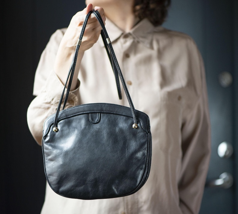 Puede incluir: Bolso de mano de cuero negro con forma redondeada y finas correas para el hombro. El bolso tiene detalles dorados en los laterales y un peque&ntilde;o cierre de solapa. La persona sostiene el bolso frente a un fondo neutro.