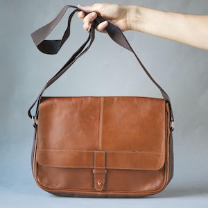 May include: A brown leather messenger bag with a flap closure and adjustable shoulder strap. The bag has a rectangular shape with a front pocket and a buckle closure. The strap is dark brown and the bag is held up by a hand.