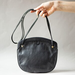 Puede incluir: Un bolso de hombro de cuero negro con forma redondeada. El bolso tiene una correa larga y fina y detalles dorados. El bolso es sostenido por una mano sobre un fondo neutro.