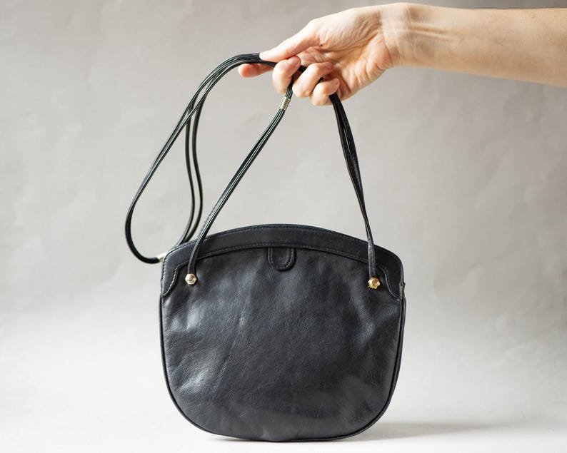 Puede incluir: Un bolso de hombro de cuero negro con forma redondeada. El bolso tiene una correa larga y fina y detalles dorados. El bolso es sostenido por una mano sobre un fondo neutro.
