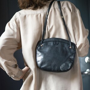 Puede incluir: Un bolso de hombro de cuero negro con forma curva y correa larga. El bolso tiene detalles dorados y cierre de solapa. Se lleva sobre una camisa beige.