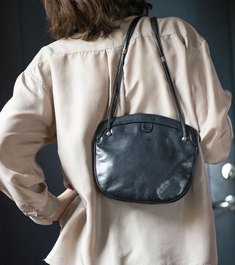 Puede incluir: Un bolso de hombro de cuero negro con forma curva y correa larga. El bolso tiene detalles dorados y cierre de solapa. Se lleva sobre una camisa beige.
