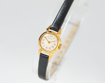 Peut inclure: Une montre-bracelet dorée avec un cadran rond blanc comportant des chiffres arabes noirs et le texte "BOMR" imprimé sous la position douze heures. La montre possède un bracelet fin en cuir noir et une couronne dorée sur le côté droit.