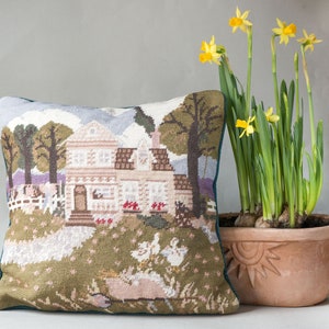 May include: A green and brown needlepoint pillow with a scene of a house, trees, and ducks. The pillow is sitting on a table next to a terracotta pot with yellow daffodils.