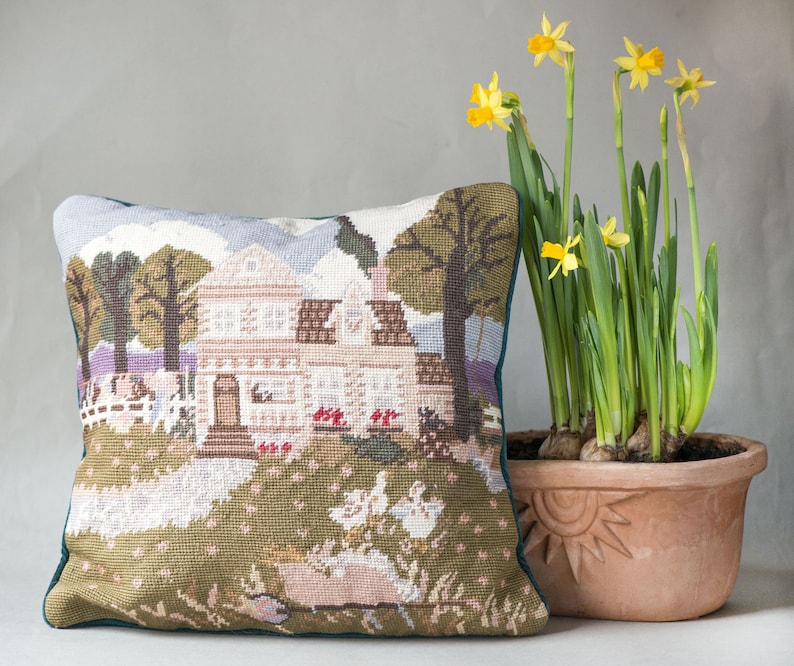 May include: A green and brown needlepoint pillow with a scene of a house, trees, and ducks. The pillow is sitting on a table next to a terracotta pot with yellow daffodils.