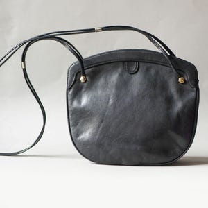 Puede incluir: Un bolso bandolera de cuero negro con la parte inferior redondeada y una correa larga y fina. El bolso tiene una parte superior curvada y peque&ntilde;os detalles dorados. La correa es fina y parece ajustable.
