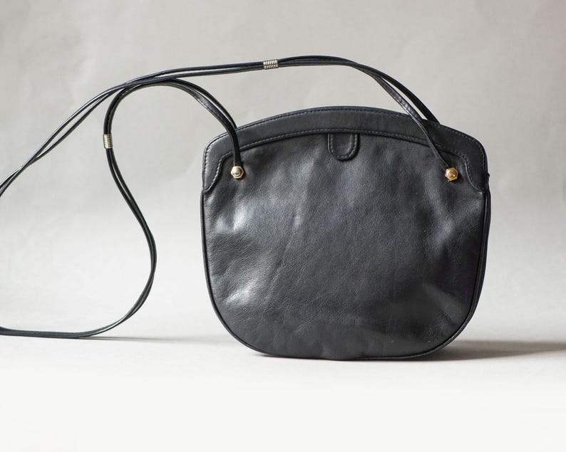 Puede incluir: Un bolso bandolera de cuero negro con la parte inferior redondeada y una correa larga y fina. El bolso tiene una parte superior curvada y peque&ntilde;os detalles dorados. La correa es fina y parece ajustable.