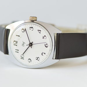 May include: A silver wristwatch with a white face and black hands. The watch has black Arabic numerals and a black leather band.