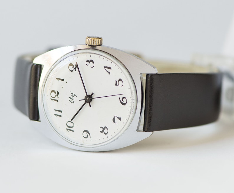 May include: A silver wristwatch with a white face and black hands. The watch has black Arabic numerals and a black leather band.