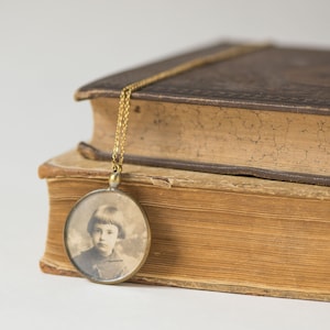 Puede incluir: Un collar de cadena dorada con un medallón redondo que presenta una foto en blanco y negro de una persona joven. El collar está apoyado sobre dos libros antiguos.