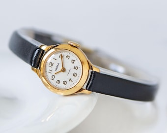 Peut inclure: Une montre-bracelet dorée avec un cadran blanc rond affichant des chiffres arabes noirs et le texte cyrillique "ЧАЙКА". La montre est dotée d'un bracelet fin en cuir noir et d'une petite couronne dorée sur le côté pour le réglage de l'heure.
