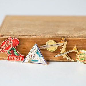 May include: Five enamel pins featuring various logos. The pins are on a brown book with a gold spine. The pins include a Coca-Cola logo, a Chippie ice cream logo, a Hapag-Lloyd Boeing 737-800 logo, a Boeing 737-800 logo, and a "Fave" logo.
