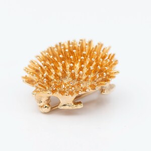 May include: A gold-toned brooch in the shape of a hedgehog. The hedgehog is made of metal and has a detailed design with many small spikes.