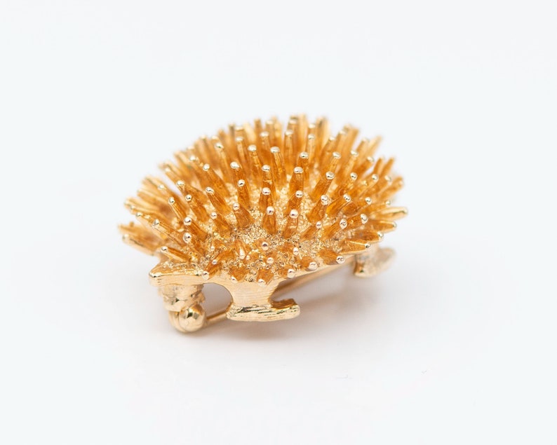 May include: A gold-toned brooch in the shape of a hedgehog. The hedgehog is made of metal and has a detailed design with many small spikes.
