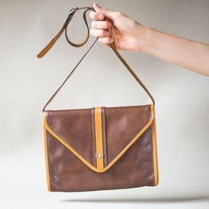 Puede incluir: Un bolso de hombro de cuero marrón con ribete de cuero beige y cierre de solapa. El bolso tiene una hebilla dorada y un pequeño logotipo dorado en la parte delantera.