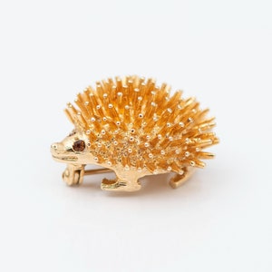 May include: A gold-toned brooch in the shape of a hedgehog with brown eyes. The hedgehog is detailed with textured fur and a curled tail.