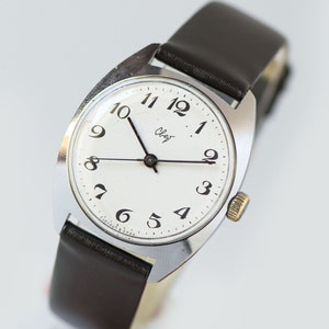 May include: A silver wristwatch with a white face and black numbers. The watch has a brown leather band and says "Слава" on the face.
