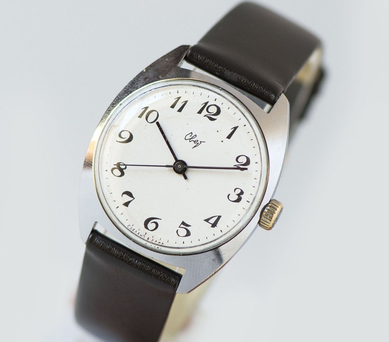 May include: A silver wristwatch with a white face and black numbers. The watch has a brown leather band and says "Слава" on the face.
